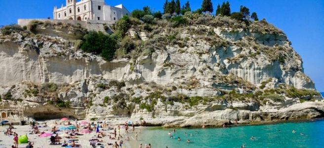 A white castle in the city of Tropea, Calabria, sits atop a sheer cliff of white stone. There is a small park behind the castle and at the foot of the cliff a beach with people enjoying the sun and sea.